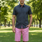 Charger l'image dans la galerie, Man wearing a black geometric  patterned shirt with pink dots and pink shorts standing in a park.
