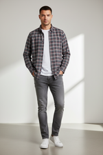 Load image into Gallery viewer, A man wearing a gray-blue-and-red plaid shirt, a white T-shirt, gray jeans, and white sneakers in a minimalist setting.
