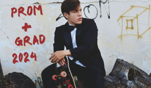 Stylish young guy in classy tuxedo sitting on log with skateboard in hand