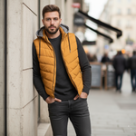 Load image into Gallery viewer, Man wearing a mustard yellow  puffer vest with removable grey hood leaning against a wall in an outdoor setting.
