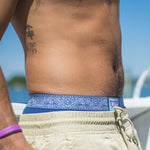 Charger l'image dans la galerie, A man  wearing blue underwear with blue and white patterned waistband with a visible brand logo on the waistband, standing by a body of water with a boat in the background.

